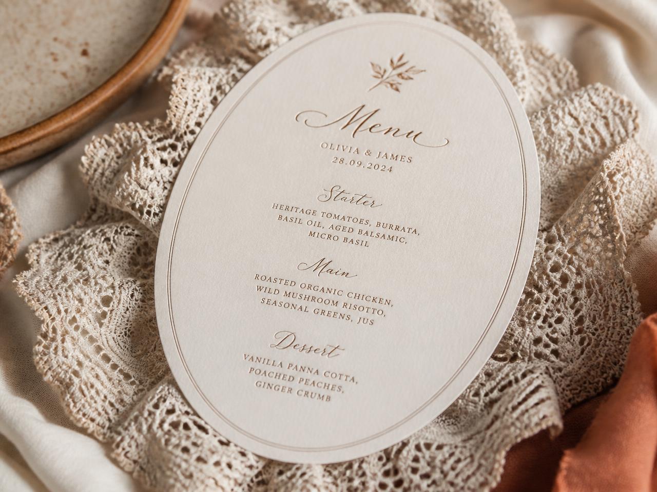 Oval engraved wedding menu on cream cotton paper resting on ivory lace